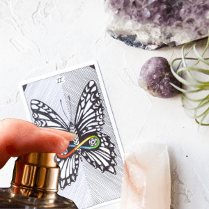 Mano rociando perfume sobre carta de tarot de mariposa junto a cristales de cuarzo